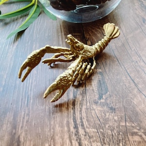 Könnte beinhalten: Eine goldfarbene Metallfigur eines Hummers. Der Hummer ist mit ausgestreckten Scheren und eingerolltem Schwanz posiert. Die Figur steht auf einer Holzoberfläche.