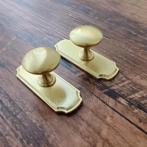 以下が含まれることがあります： 丸いノブのデザインの真鍮色のキャビネットプルが2つ。各プルには、装飾的なエッジが付いた長方形のバックプレートが付いています。ハードウェアは木製の表面に表示されます。