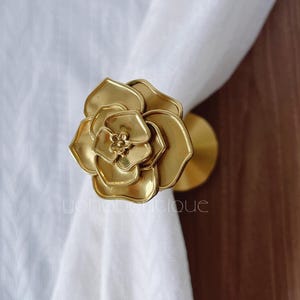 May include: A gold-coloured metal curtain tieback in the shape of a rose. The rose has multiple petals and a detailed centre. The tieback is holding a white curtain. The design is elegant and decorative.