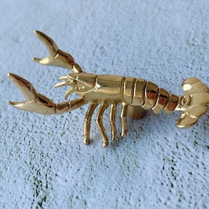 May include: A gold-colored lobster figurine with detailed claws, legs, and segmented body. The crustacean is posed on a light blue and white textured surface. This decorative item could be used as a cabinet pull or a decorative accent.