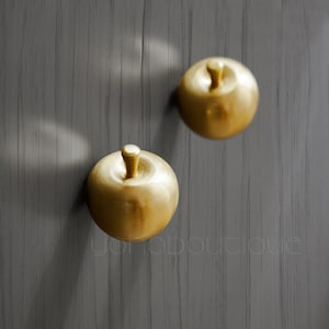 May include: Two gold apple-shaped door knobs on a grey wood background.