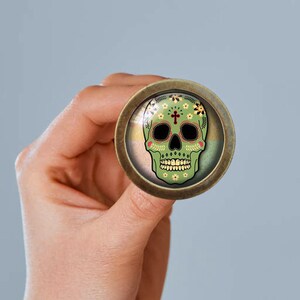 May include: A green and gold sugar skull with floral details and a cross on the forehead, displayed on a round, bronze-coloured pin.