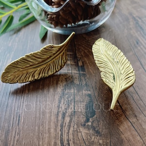 May include: Two gold-colored metal feather-shaped decorative pieces on a wooden surface.