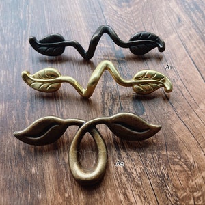May include: Three decorative cabinet pulls with a leaf design. The pulls are made of metal and have a gold, bronze, and black finish.