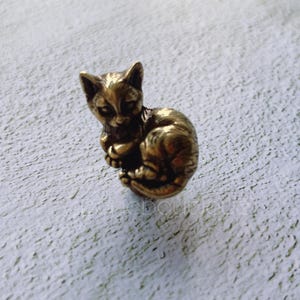 Może przedstawiać: Mała, złota figurka kota wykonana z metalu. Kot jest zwinięty w pozycji do spania, z głową opartą na łapach.