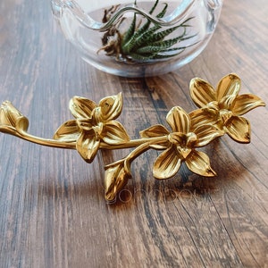 May include: A gold-toned metal orchid flower design with three blossoms. The flowers are detailed with intricate carvings and have a delicate, feminine aesthetic.