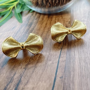 Könnte beinhalten: Zwei goldfarbene Schubladenknöpfe in Form einer Schleife.