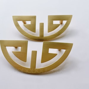 May include: Two gold-toned metal cabinet pulls with a geometric design. The pulls are shaped like half circles with a cutout pattern.