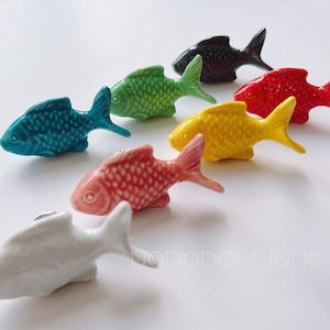 Peut inclure: Sept figurines de poissons en céramique de différentes couleurs, dont le rouge, le jaune, le vert, le bleu, le rose, le noir et le blanc. Les poissons sont tous tournés vers la droite de l'image et ont un design 3D.