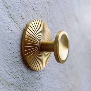 Puede incluir: Un gancho de metal dorado con un diseño de sol en la base. El gancho está montado en una pared blanca.
