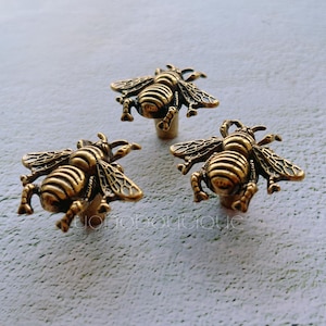 Pode incluir: Três puxadores de armário em forma de abelha, cor bronze. Cada puxador apresenta asas e segmentos corporais detalhados, com uma haste pequena para fixação. Os puxadores estão dispostos sobre uma superfície cinza claro, mostrando seu design intrincado.