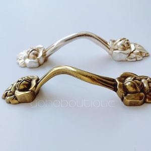 Op de afbeelding: Twee sierlijke kastgrepen, een zilveren en een messing, met een bloemmotief. De grepen zijn gebogen en hebben een decoratief, reliëf bloemmotief.