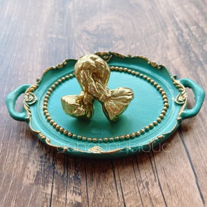 Könnte beinhalten: Ein goldfarbenes Dekorationsobjekt in Form eines Knotens liegt auf einem türkisfarbenen Tablett mit goldenen Akzenten und einem Perlenrand.