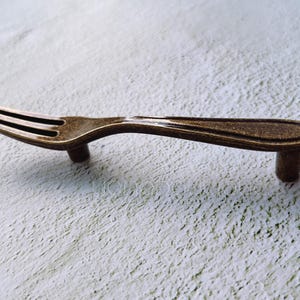 Könnte beinhalten: Ein bronzefarbener, gabelförmiger Schrankgriff mit schlichtem Design.