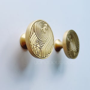 Pode incluir: Dois ganchos de metal dourado com um design circular que apresenta um padrão em espiral. Os ganchos estão montados em uma parede branca.