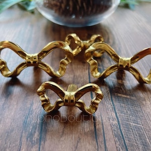 May include: Four gold-colored bow-shaped cabinet pulls. The pulls are made of metal and have a detailed, textured design. The bows are arranged on a dark wooden surface, with a glass bowl and greenery in the background.