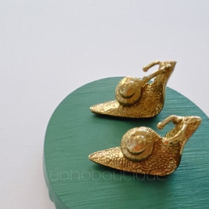 Puede incluir: Dos pendientes dorados con forma de caracol. Los pendientes están sobre una superficie verde. Los caracoles tienen cuerpos texturizados y conchas detalladas. Los pendientes son probablemente de metal y tienen un acabado brillante.