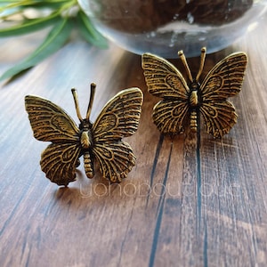 May include: Two antique brass butterfly-shaped decorative objects. The butterflies have detailed wing patterns and antennae. They are displayed on a wooden surface. The word "yananboutique" is visible in the image.