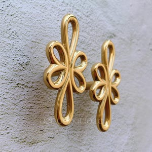 May include: Two gold-toned metal decorative hooks in the shape of a stylized flower. The hooks are mounted on a white wall.