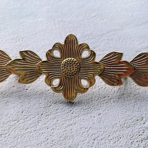 Op de afbeelding: Een decoratief bloemaccentstuk van messing. Het stuk heeft een centraal bloemontwerp met een getextureerd midden en bloembladachtige elementen die naar buiten uitsteken. Het messing heeft een gepolijste afwerking.