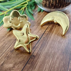 Op de afbeelding: Drie gouden metalen ornamenten met lachende gezichten. De ornamenten zijn gevormd als een wolk, een ster en een halve maan.