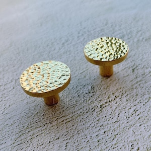 以下が含まれることがあります： ハンマーで叩いたような質感の、丸い金色のキャビネットノブが2つ