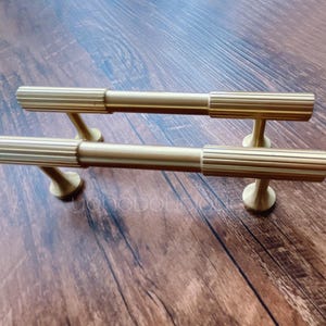 Puede incluir: Dos tiradores de armario dorados con un diseño acanalado. Los tiradores están montados en una superficie de madera.