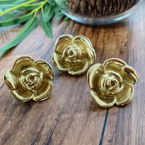 Könnte beinhalten: Drei goldfarbene, rosenförmige Schrankknöpfe. Die Knöpfe sind aus Metall und haben eine glänzende Oberfläche. Die Knöpfe sind auf einer Holzoberfläche angeordnet.