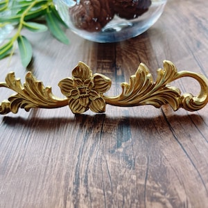 May include: Gold-toned metal drawer pull with a floral design. The pull features a stylised flower in the centre with intricate leaf details on either side.