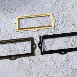 May include: Three rectangular label holders in brass, bronze, and black. Each has a central opening for a label and two screw holes on each side. The holders are arranged on a textured, light gray surface.