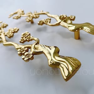May include: Two gold-toned cabinet pulls shaped like stylized trees with blossoms. The pulls are detailed and have a smooth, polished finish.