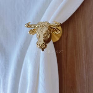 May include: A gold-coloured bull head curtain tieback. The bull head is highly detailed with horns, ears, and facial features. The tieback is attached to a round base. The text "yohoboutique" is visible at the bottom.