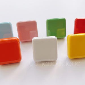 以下が含まれることがあります： さまざまな色（薄い青、ピンク、緑、赤、黄、白、オレンジ）の正方形のセラミック製キャビネットノブが7つあります。