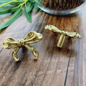 Könnte beinhalten: Zwei goldfarbene, schleifenförmige Schrankknöpfe. Die Knöpfe sind aus Metall und haben eine glänzende Oberfläche. Die Schleifen haben detaillierte Kurven und ein realistisches Aussehen. Die Knöpfe sind für Möbel und Heimdekoration bestimmt.