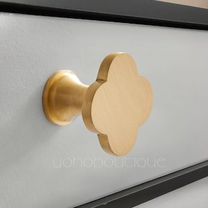 Peut inclure: Une poignée de tiroir en métal doré avec un motif de trèfle à quatre feuilles. La poignée est montée sur une surface blanche.