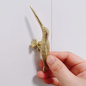 May include: A gold-colored hummingbird-shaped cabinet pull. The pull is highly detailed, with textured feathers and a long, slender beak. The hummingbird is perched on a small branch, adding a touch of nature-inspired elegance to any cabinet or drawer.