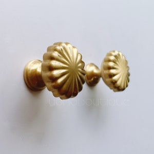 以下が含まれることがあります： 白い壁に取り付けられた、装飾的な貝殻模様の金色のキャビネットノブが2つあります