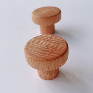 Könnte beinhalten: Zwei Holzknöpfe mit runder Oberseite und zylindrischem Fuß. Die Knöpfe sind aus hellbraunem Holz mit natürlicher Maserung.