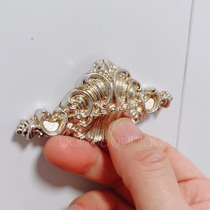 May include: A silver and gold decorative drawer pull with a detailed ornate design. The pull is shaped like a wave or a shell.