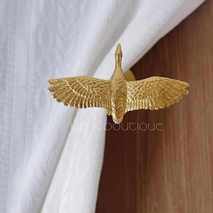 Peut inclure: Un embrasse-rideau en métal doré en forme d'oie en vol. L'oie a des ailes déployées et des plumes détaillées. L'embrasse-rideau est monté sur un rideau blanc, avec le nom de la boutique "iohoboutique" visible.