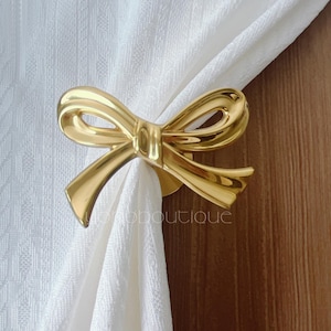 Puede incluir: Un recogedor de cortina dorado en forma de lazo. El recogedor de metal sujeta una cortina blanca. El lazo tiene un diseño detallado y está fijado a una base redonda. Un artículo decorativo.
