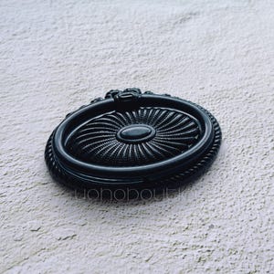 May include: Black oval drawer pull with a textured centre and rope-like edging. The hardware features a decorative design and a small, dark oval in the centre. The pull is set against a light, textured background.