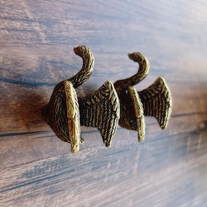 May include: Three antique brass-colored wall hooks, each featuring a stylized wing design. The hooks are mounted on a wooden surface with visible grain. The hooks are designed to hold items.