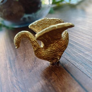 May include: A gold-colored swan-shaped ring with textured details. The ring is set on a wooden surface. The ring's design includes a curved neck and outstretched wings. The background includes a glass container with decorative elements.
