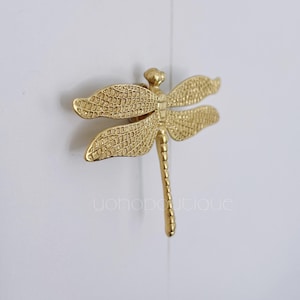 May include: A gold-toned dragonfly-shaped door knob or pull. The dragonfly has detailed wings and a long, slender body.
