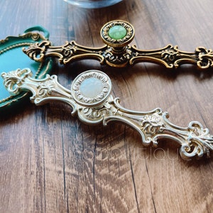 Puede incluir: Dos pomos de armario de color dorado con detalles decorativos. Un pomo tiene un centro de vidrio verde y el otro tiene un centro de vidrio blanco.