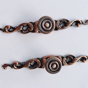 Peut inclure: Deux poignées de porte de placard en cuivre orné avec un motif de défilement décoratif et un bouton rond.