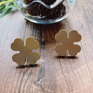 Pode incluir: Dois brincos de trevo de quatro folhas de cor dourada sobre uma superfície de madeira.