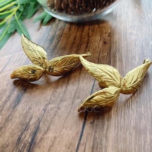 Könnte beinhalten: Zwei goldene Haarklammern in Blattform. Die Klammern sind aus Metall und haben eine glänzende Oberfläche.