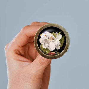 Könnte beinhalten: Ein runder, messingfarbener Knopf mit einem weißen Magnolienblüten-Design auf schwarzem Hintergrund.